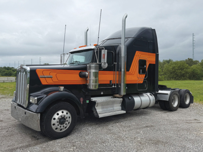 Kenworth W900L AeroCab tractor in profile showing orange and black paint, with chrome fuel tanks and dual exhaust stacks.
