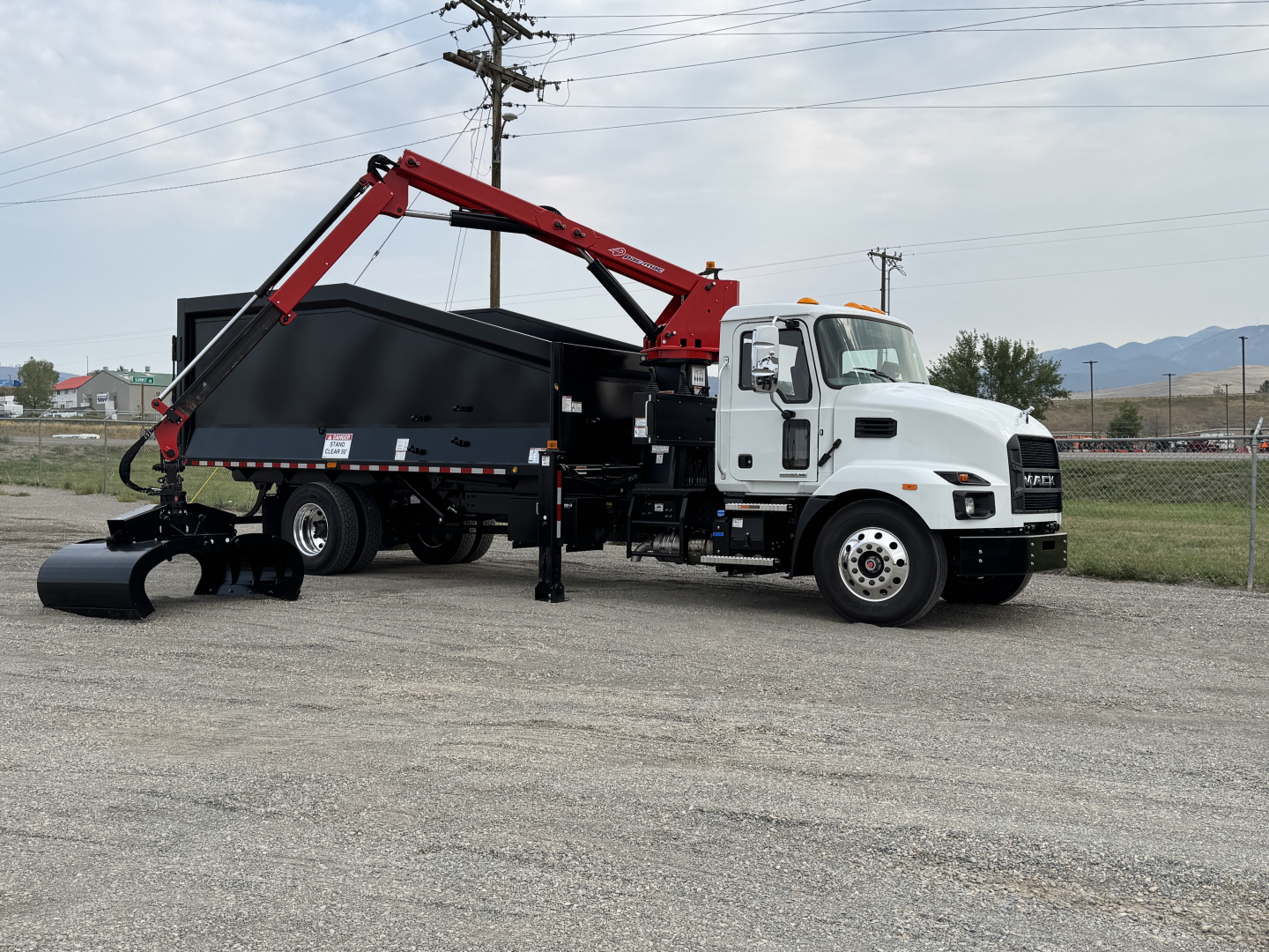 2025 Mack MD7 grapple truck with a red hydraulic arm and black dump body parked on a lot; truck cab white, warning lights on top, mountains in distance.