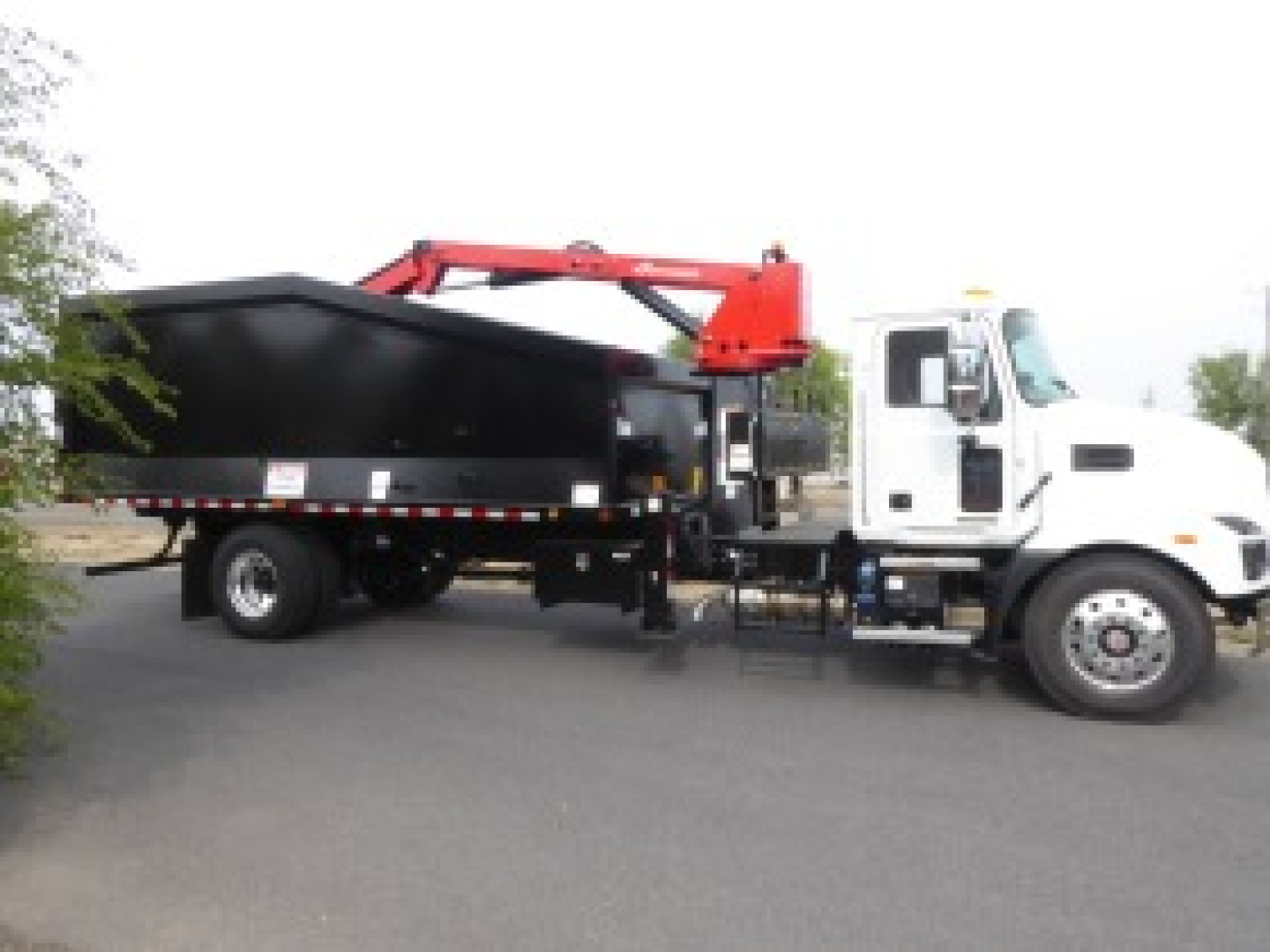 Truck cab with a large black dump bed and red hydraulic arm attached to a white Mack MD7 truck. Grapple truck setup visible.