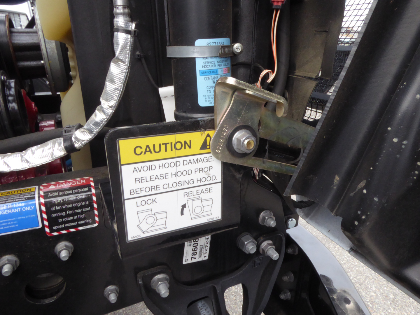 Mack MD7 truck component close-up showing caution label and hood latch hardware with warning stickers and metallic fittings.