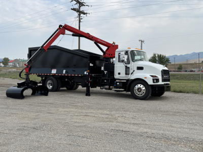 2025 Mack MD7 grapple truck with a red hydraulic arm and black dump body parked on a lot; truck cab white, warning lights on top, mountains in distance.