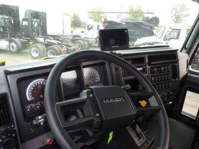 Mack MD7 cab interior showing steering wheel with MACk logo, dashboard gauges, and infotainment screen; exterior lot with other trucks visible through windshield.