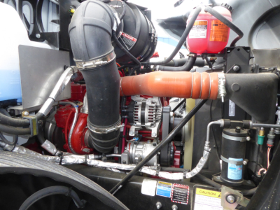 Mack MD7 engine bay showing air intake piping, alternator, orange coolant hose, red engine components, and various hoses and reservoirs.