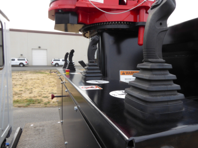 Grapple truck equipment on a Mack MD7 chassis. Visible text includes warning labels and circular decals on a black glossy surface near hydraulic mountings.