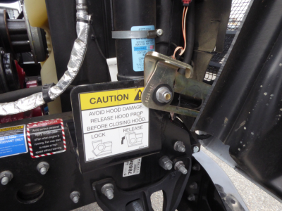 Mack MD7 truck component close-up showing caution label and hood latch hardware with warning stickers and metallic fittings.