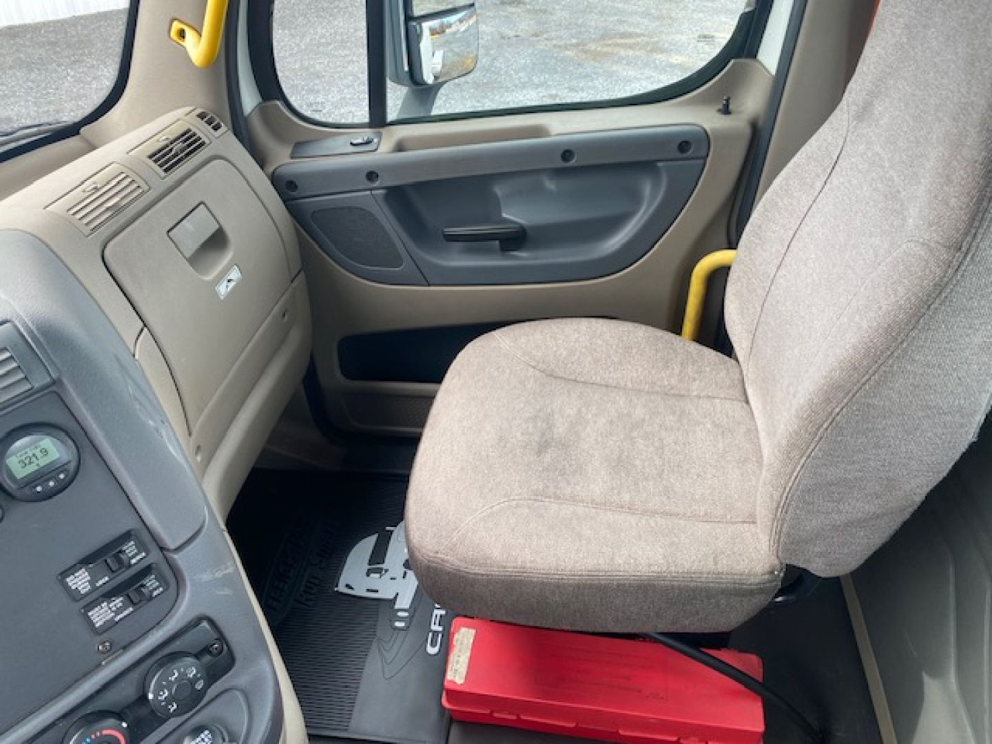Cab interior of a 2016 Freightliner Cascadia 113 daycab showing beige dashboard, steering area, and gray fabric seats with a red toolbox or item under the passenger seat.