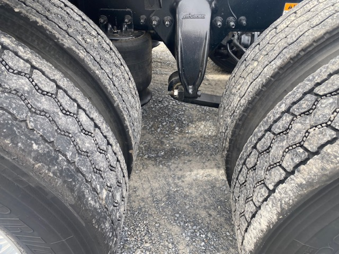 Front view of Freightliner Cascadia 113 tractor dual rear axle tires with fifth wheel hitch and driveline components visible in center, gravel ground.