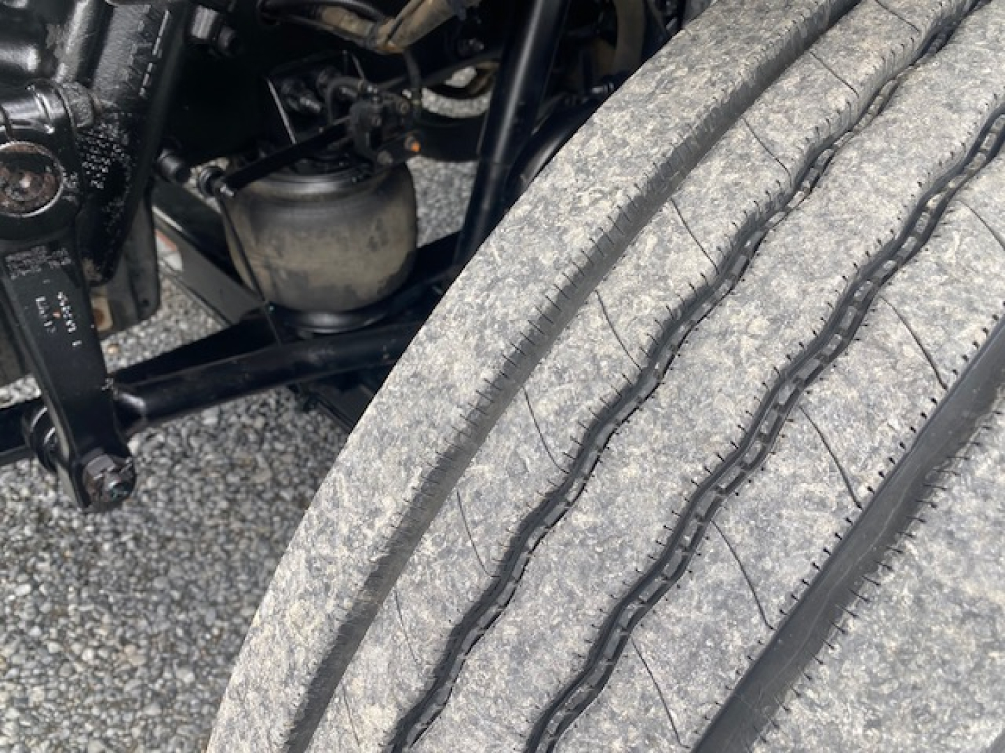 Truck front axle area with heavy-duty tire and suspension components. Visible components include a large tire tread, part of the wheel well, and off to the left engine bay elements and hoses.