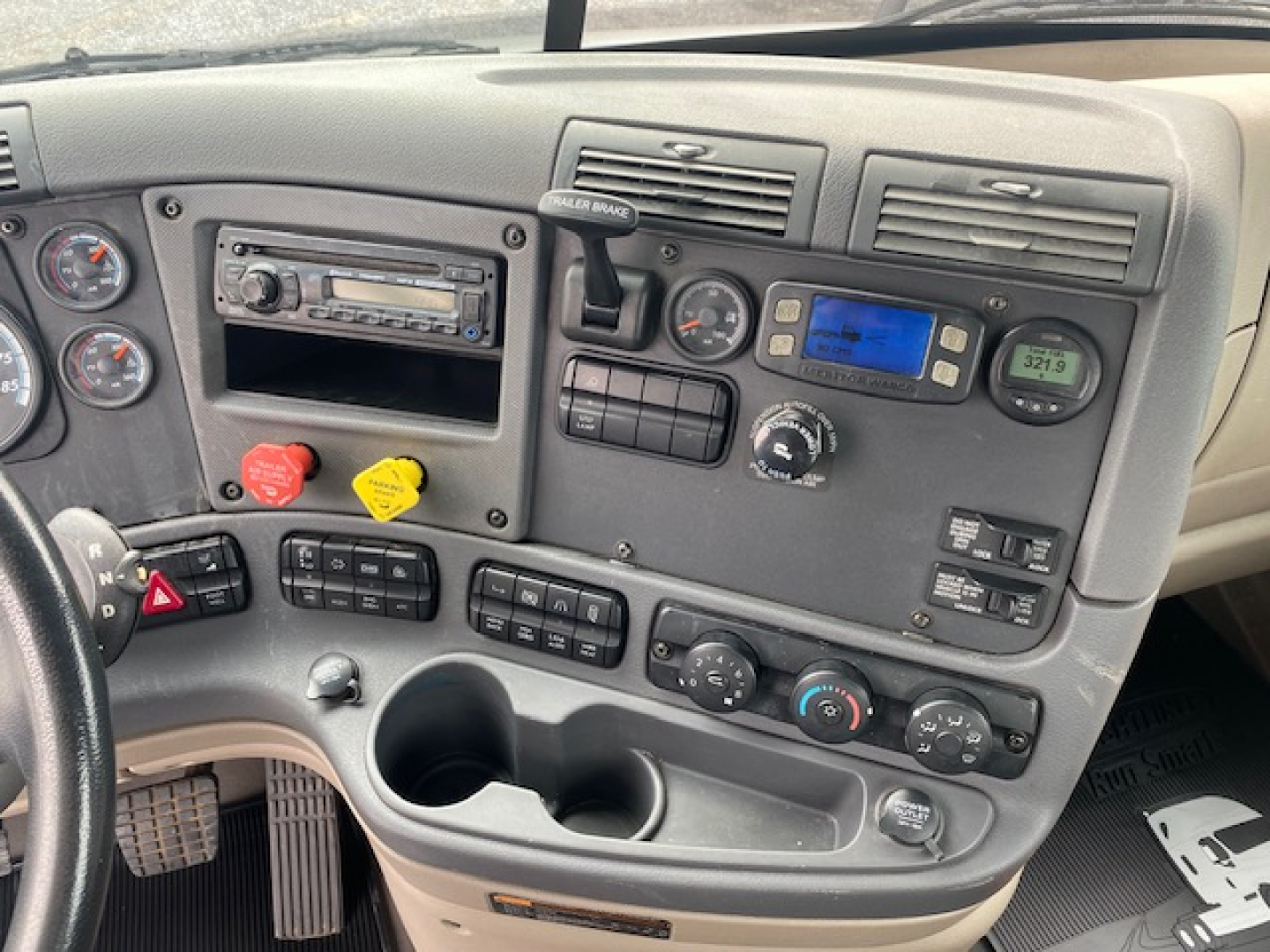 Cab interior of a Freightliner Cascadia truck showing dashboard gauges, control switches, cup holders, and stereo unit.