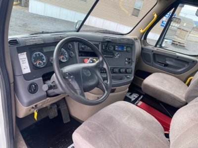 Cab interior of a 2016 Freightliner Cascadia 113, view of steering wheel, dashboard gauges, and driver seat from passenger side, with yellow grab handle and center console controls.