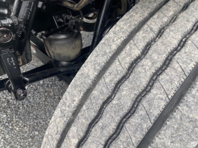 Truck front axle area with heavy-duty tire and suspension components. Visible components include a large tire tread, part of the wheel well, and off to the left engine bay elements and hoses.