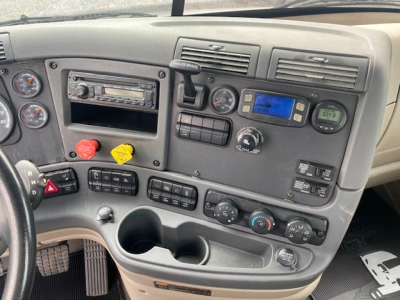 Cab interior of a Freightliner Cascadia truck showing dashboard gauges, control switches, cup holders, and stereo unit.