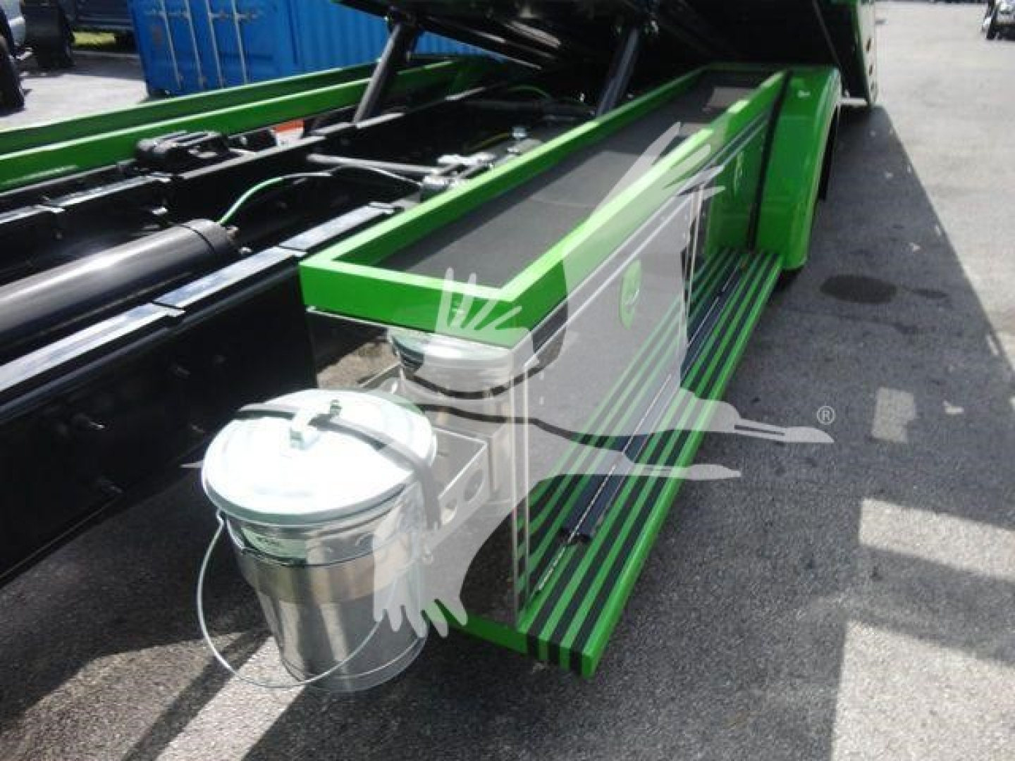 Truck bed area of a green International MV 2027 model showing dual side steps and a stainless steel bucket with handle near the wheel well.