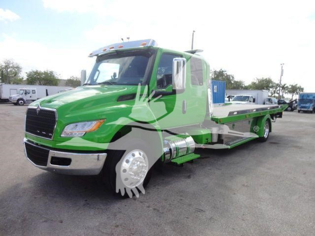International MV tow truck on site, green heavy-duty rollback with extended flatbed, chrome bumper, side fuel tank, and large wheel covers visible.