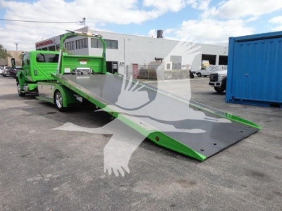 Green rollback tow truck with long tilt-bed flatbed in a parking lot, International MV chassis visible in background.