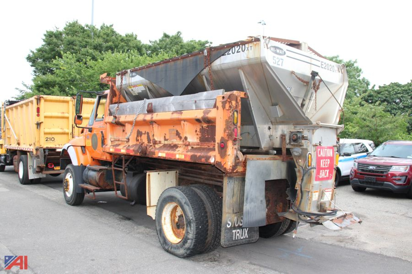 Dump truck with hopper attached, rear view showing orange bed, gray dump mechanism, and white fuel or water tank, side lettering and license plate area.