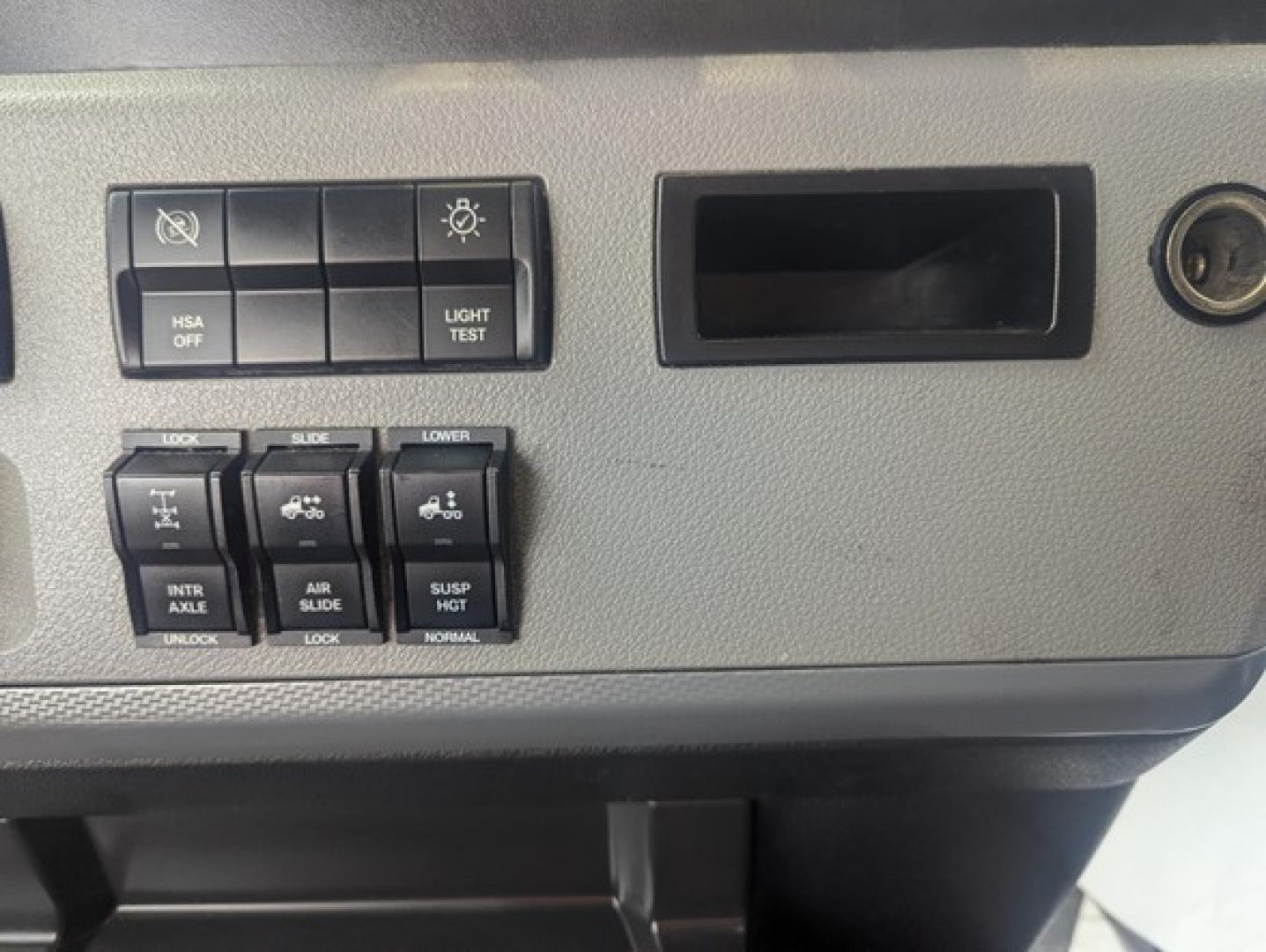 Cabin dash area of a Freightliner Cascadia showing control switches including light/test, air suspension, and differential locks, with a small storage opening.