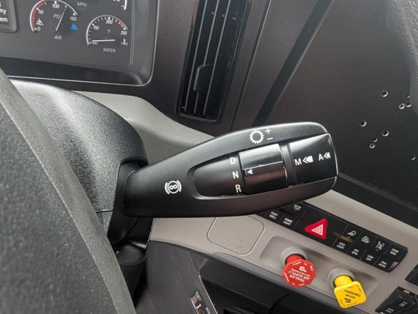 Truck interior steering column shifter and controls of a 2022 Freightliner Cascadia 126 showing gear stalk with D N R P and cruise control symbols, dashboard gauges in background, red and yellow switches.