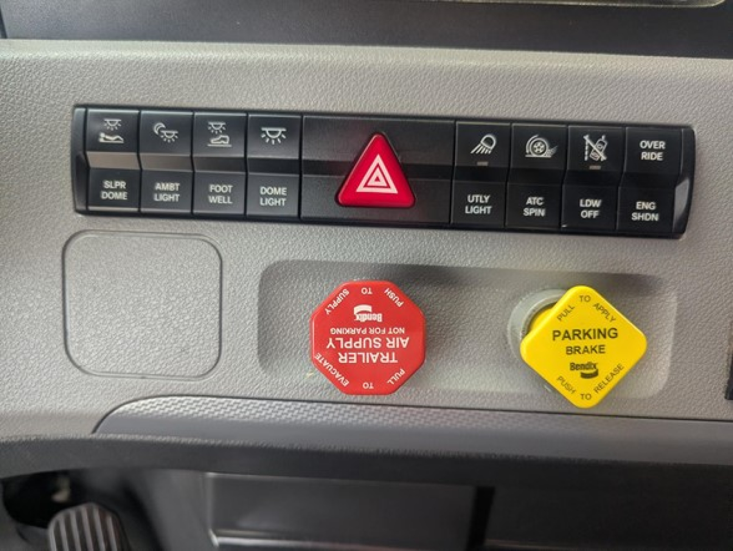Dashboard controls of a Freightliner Cascadia showing red emergency stop button and yellow parking brake knob with text; row of small labeled switches above