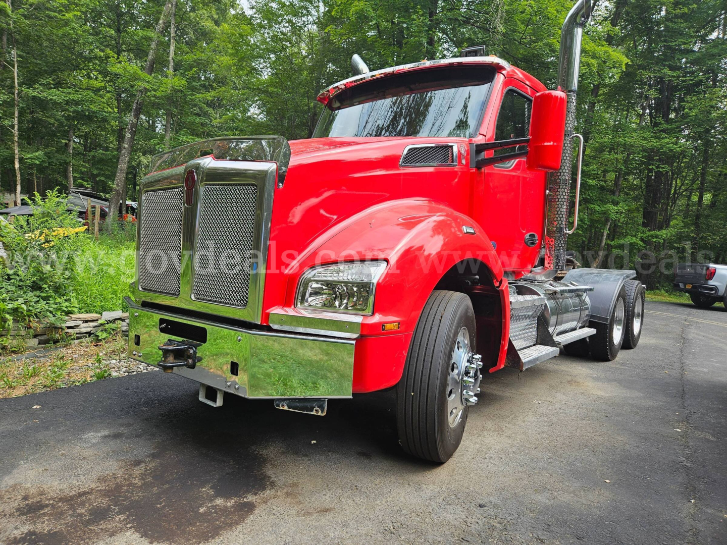 Kenworth T880 truck in red, front-left view showing chrome grille, large bumper, dual headlights, and dual rear axles with a sleeperless cab; forested background.