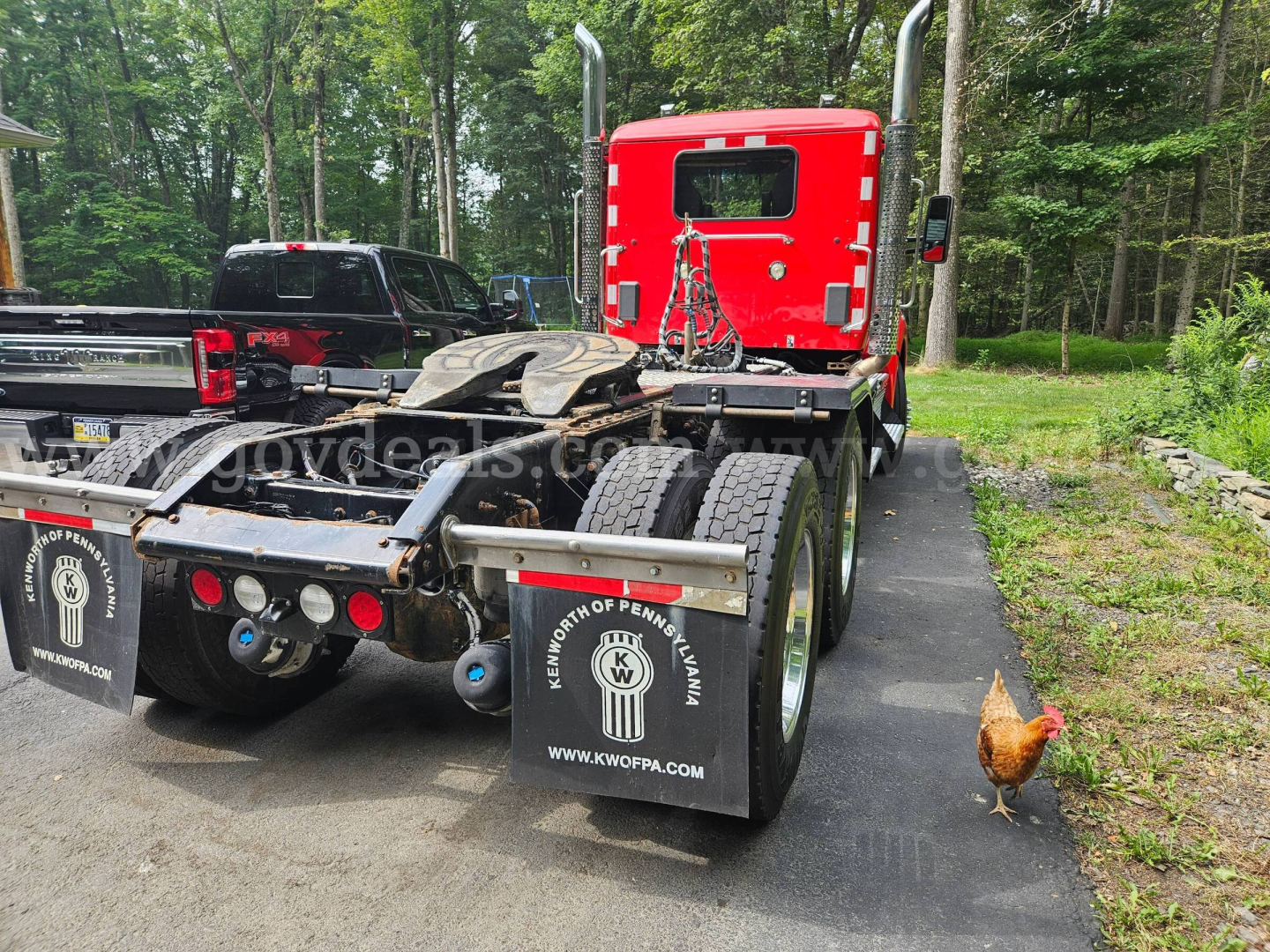 Kenworth T880 tractor truck rear view showing fifth wheel and dual rear axles, black mudflaps with KW ... logo, red cab, small chicken on right side.