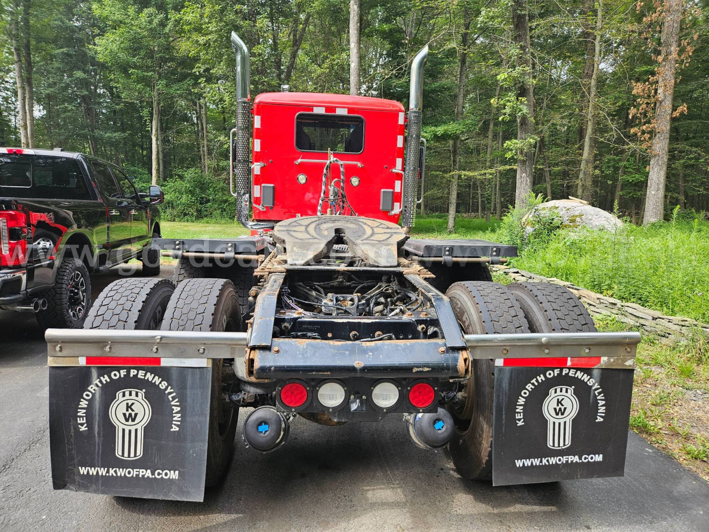 Kenworth truck rear view showing red cab, dual rear axles, mud flaps with KW logo, and chassis with fifth wheel.