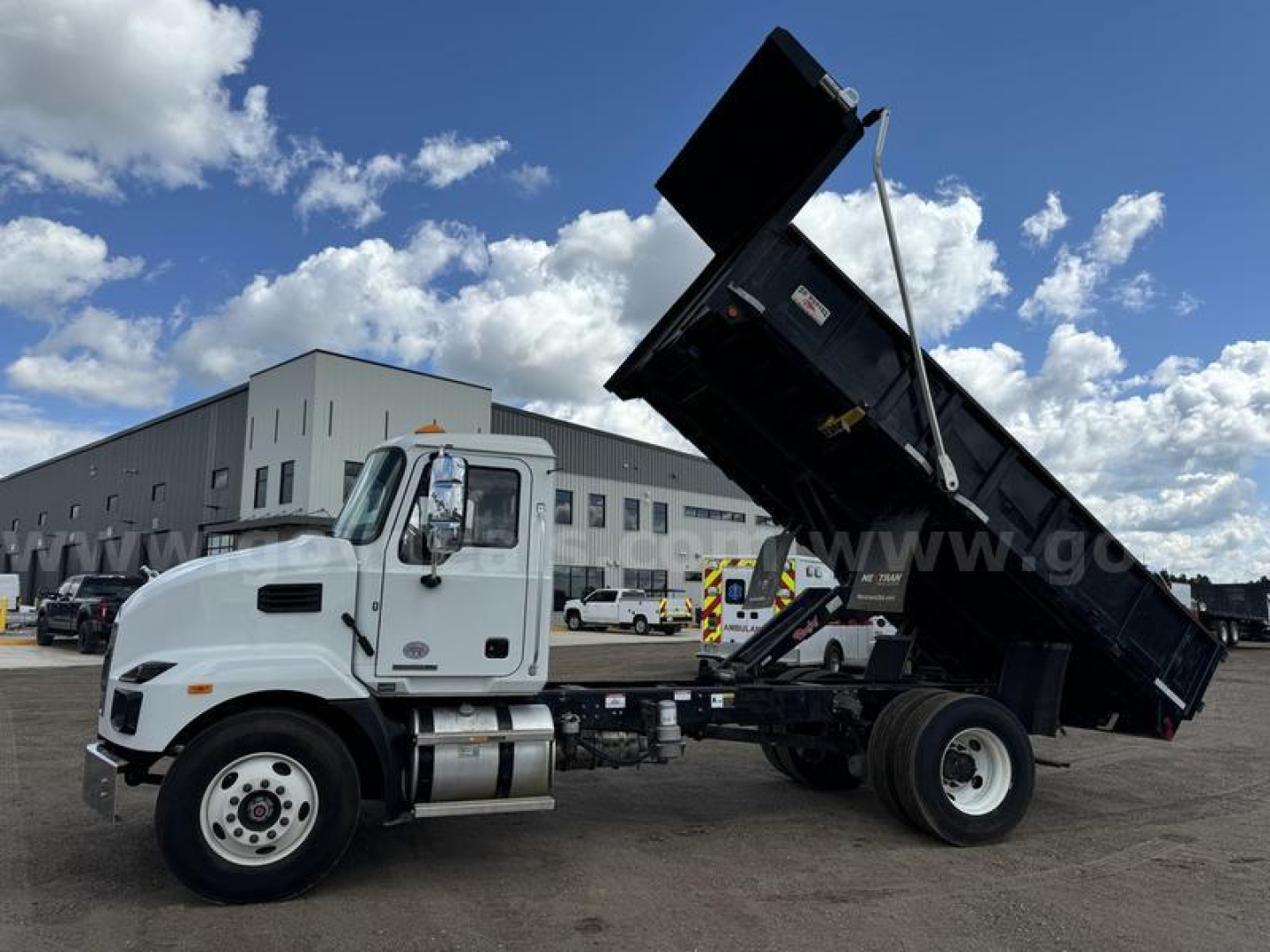 Dump truck in-service showing Mack MD cab and raised dump bed with hydraulic lift. Fleet yard background with blue sky and clouds.