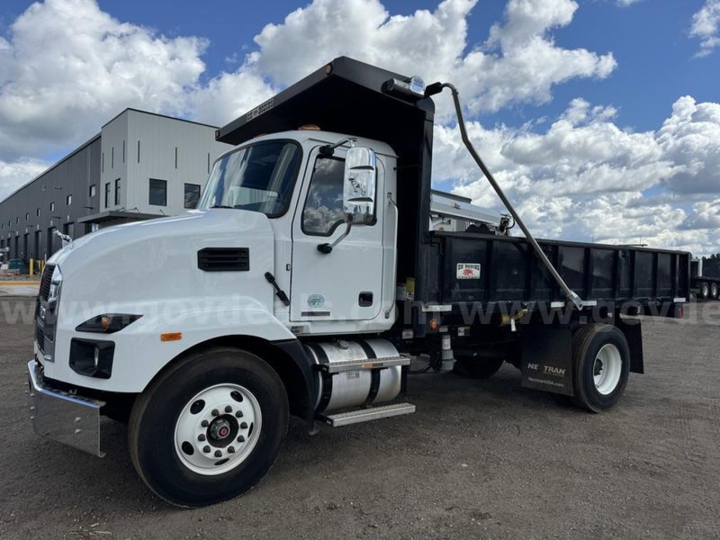 Dump truck cab and bed with black dump body, Mack MD logo visible on grille and side, front tire, rear hydraulic dump system, blue sky with clouds.