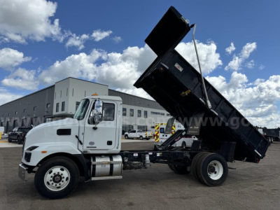 Dump truck in-service showing Mack MD cab and raised dump bed with hydraulic lift. Fleet yard background with blue sky and clouds.