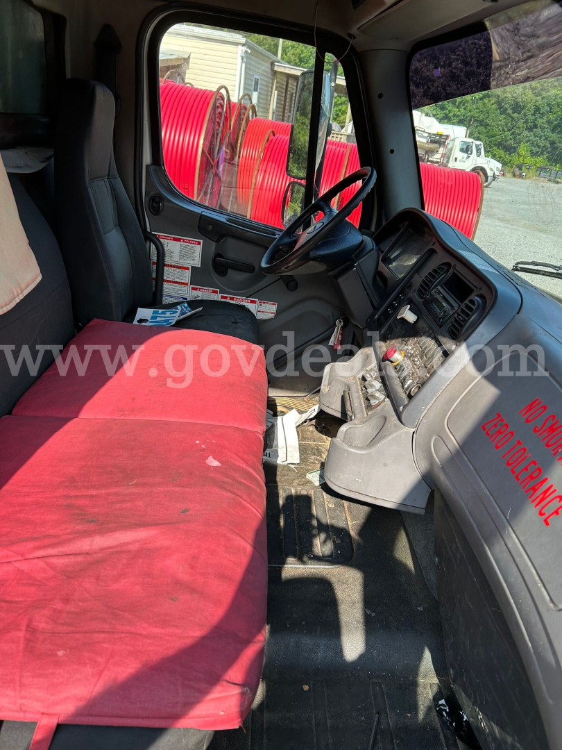Cab interior of a Freightliner M2 106 truck showing red bench seat, steering wheel, dashboard with controls, and windshield; visible red hoses outside.