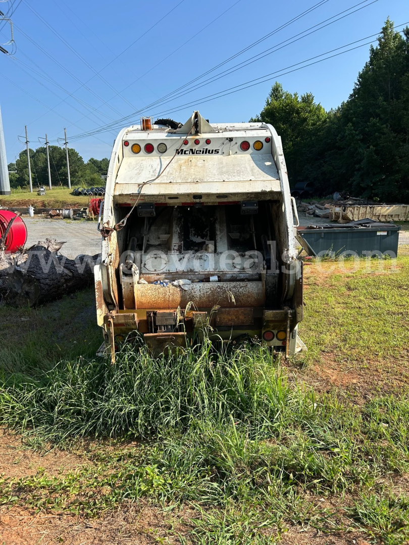 Rear view of a white Freightliner M2 106 dump truck body showing McNeilus bed and tailgate; rust and dirt on the debris chute; grass surrounding the hitch area.