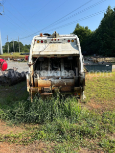Rear view of a white Freightliner M2 106 dump truck body showing McNeilus bed and tailgate; rust and dirt on the debris chute; grass surrounding the hitch area.