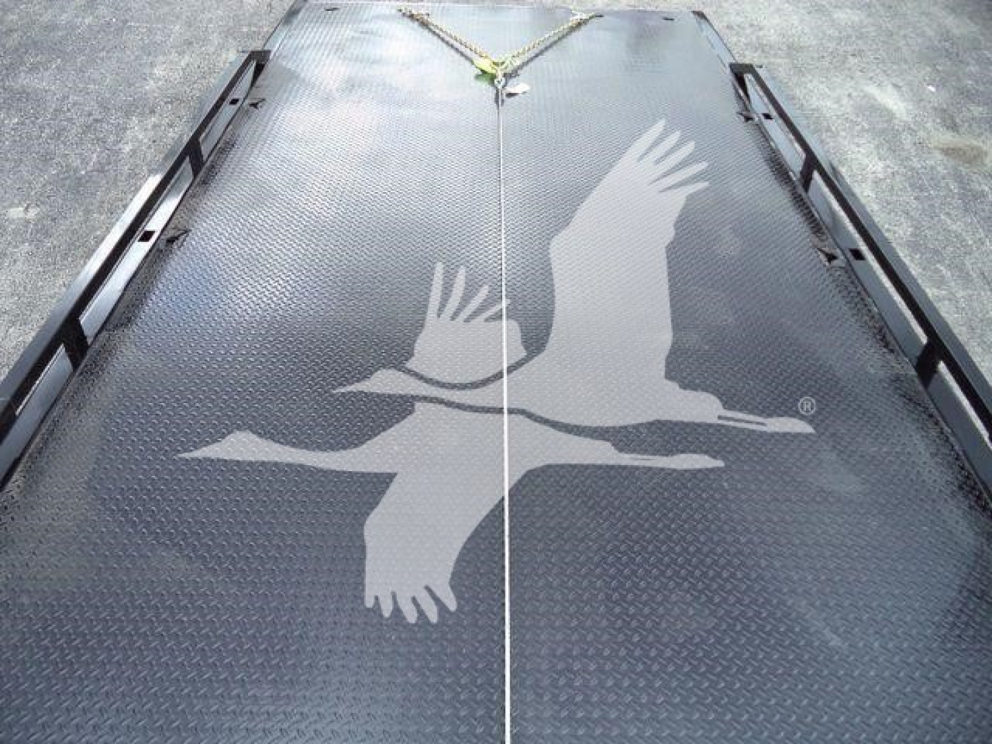 Truck bed with black diamond plate surface and a white crane logo, tie-down hooks at edges, viewed from rear, part of a rollback truck.
