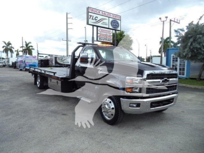 Truck bay showing a Chevrolet Silverado 6500HD rollback tow truck with a flatbed. Visible text on signs in background reads TLC Truck Sales Center and Jerrad? etc. No other legible text on components.