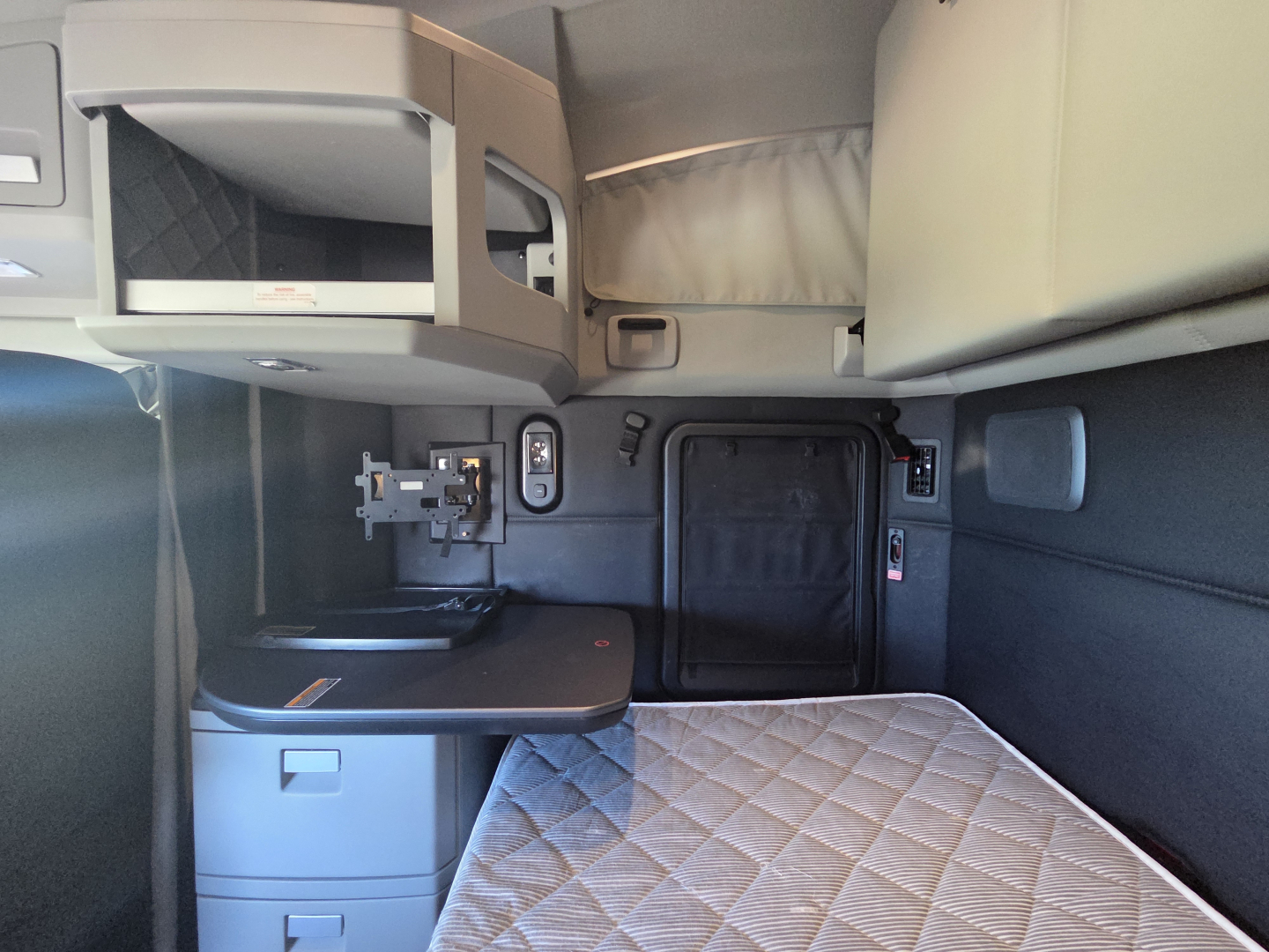 Kenworth T680 cab interior showing sleeper area with storage shelf, fold-out desk, and quilted mattress in the sleeping berth.