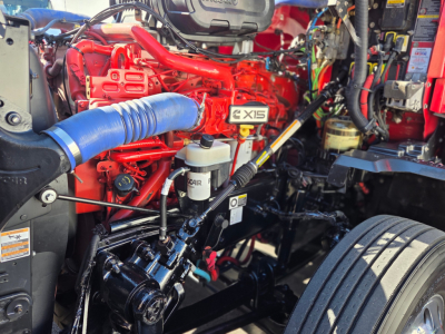 Kenworth T680 engine bay showing EXIS badge and surrounding hoses, wiring, and components in red and black with a blue coolant hose and a white fluid reservoir.