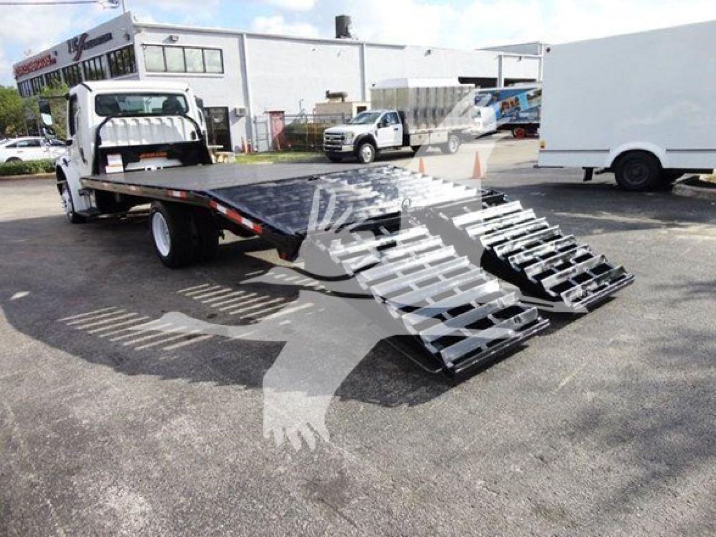 Flatbed tow truck with a ramped loading bed in an outdoor lot; Freightliner M2 106 style flatbed with gooseneck and wheel ramps visible.
