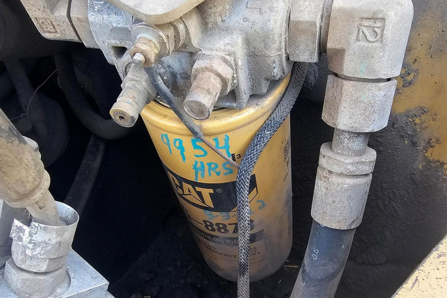 Caterpillar 902 wheel loader fluid reservoir and hydraulic hoses with visible stamp: 'CAT' and serial-like markings on the yellow filter canister. Various metal fittings and hoses surround.