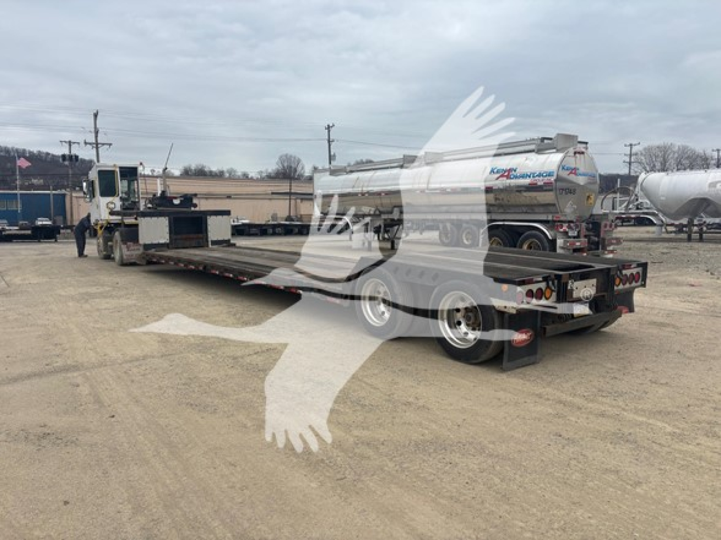 Trailer showing a long flatbed with a double-drop expandable section and a cylindrical tanker on a white chassis, rear view with lights and dual axles.