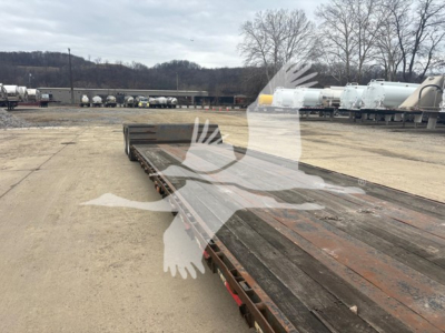 Trailer deck view of a double drop expandable trailer with weathered steel and rust patina, long flatbed surface, exposed frame, and distant equipment in lot.