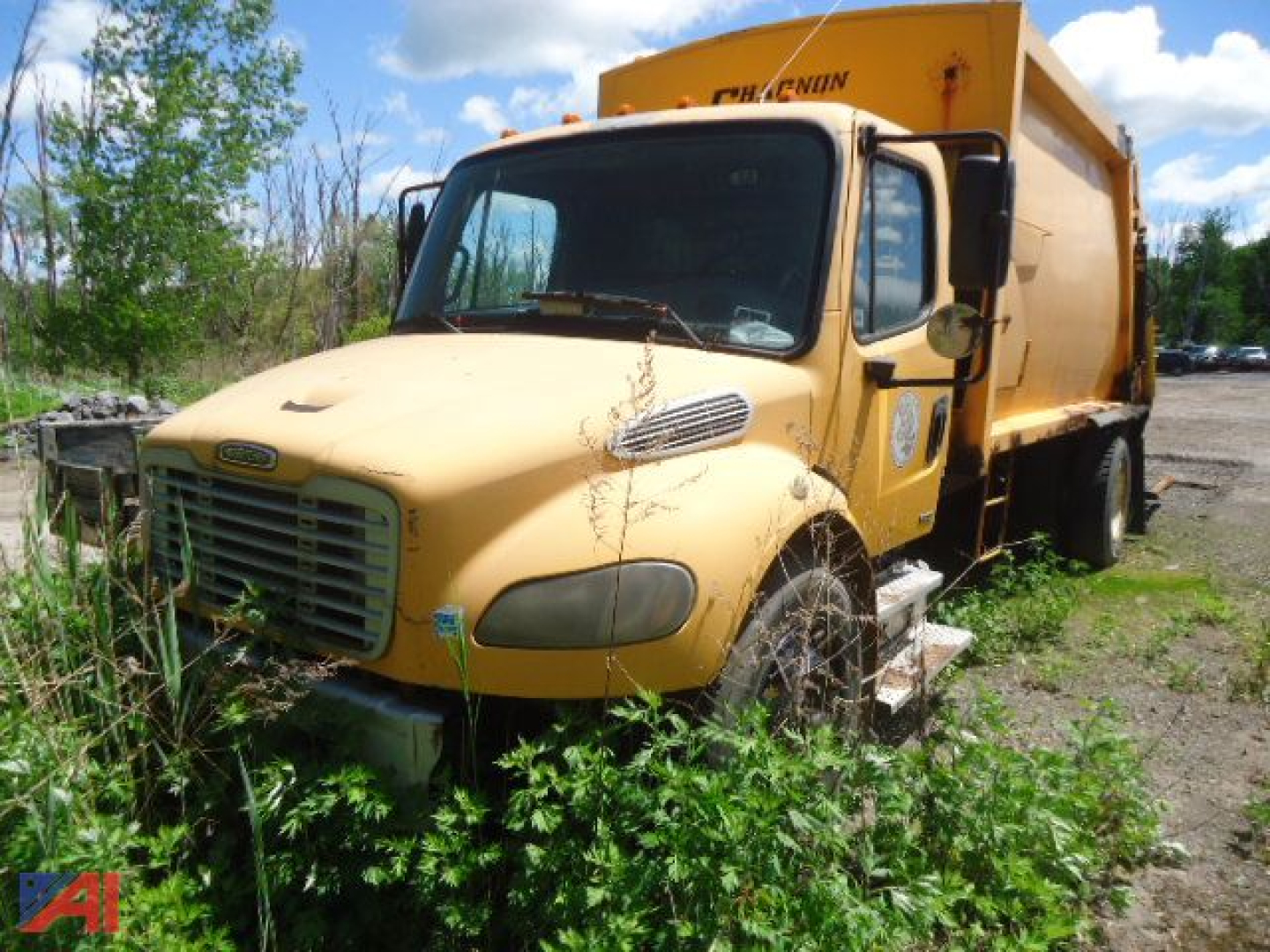 Medium shot of a yellow 2005 Freightliner M2 106 refuse truck with a large rear hopper, parked among tall weeds; front grille, bumper, and wheel wells visible.