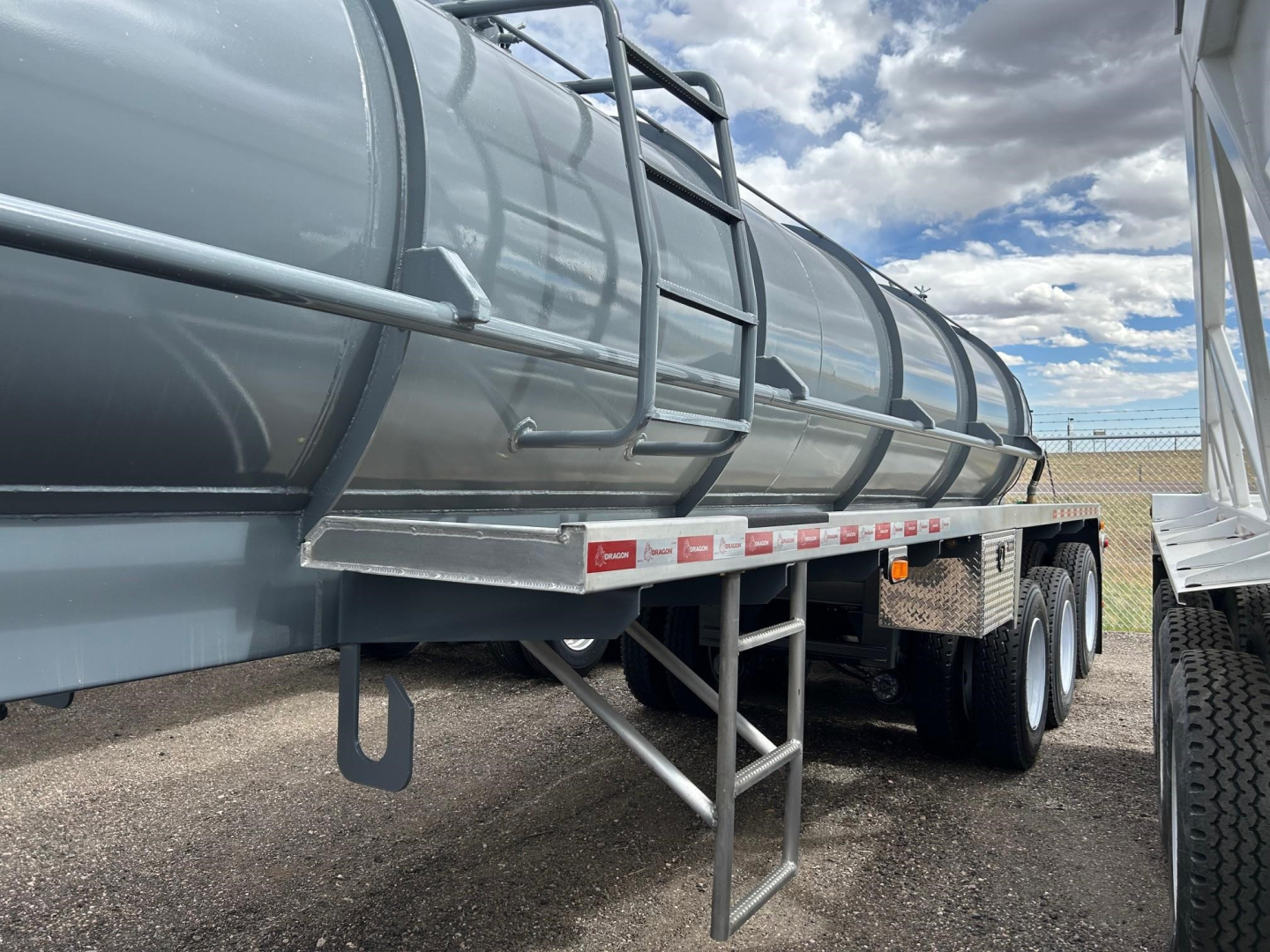 Tank trailer with cylindrical steel tank on triple axles. Side ladder and steps beneath, with small red-and-white reflectors along the frame. Clear blue sky and fence in background.