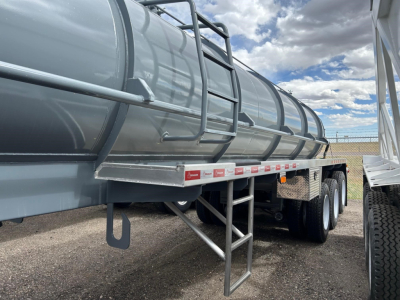 Tank trailer with cylindrical steel tank on triple axles. Side ladder and steps beneath, with small red-and-white reflectors along the frame. Clear blue sky and fence in background.