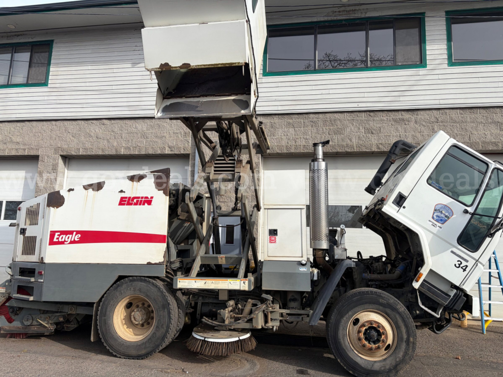 Close-up of a street-sweeper truck labeled Eagle Elgin, with a damaged front cab and elevated hopper showing mechanical broom. Side view includes dirty tires and metal framework.