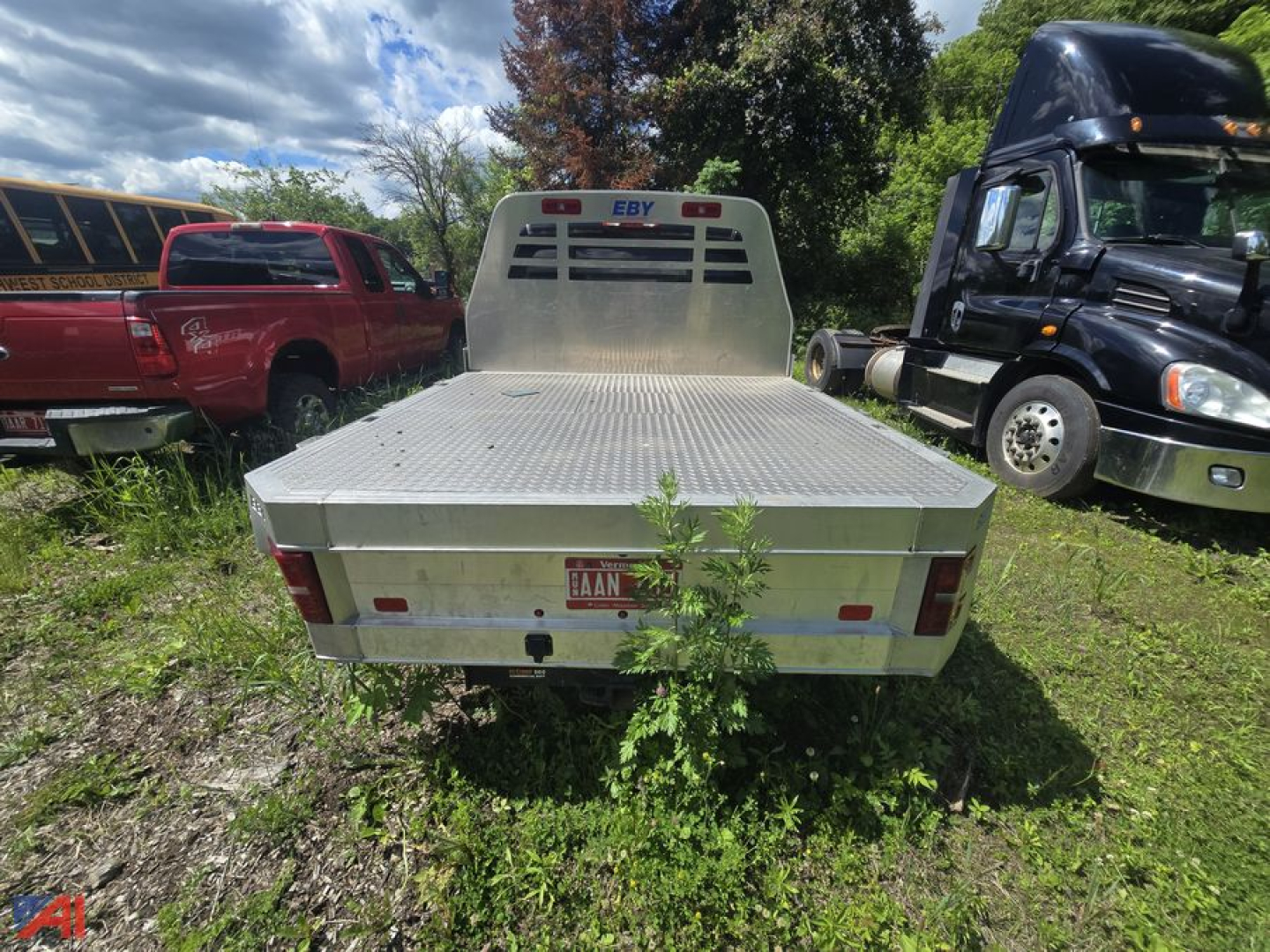 Flatbed rear view of a 2005 Chevrolet Silverado 2500HD with aluminum flatbed and vertical cab guard; surrounding vehicles include a red pickup and a black semi-truck.
