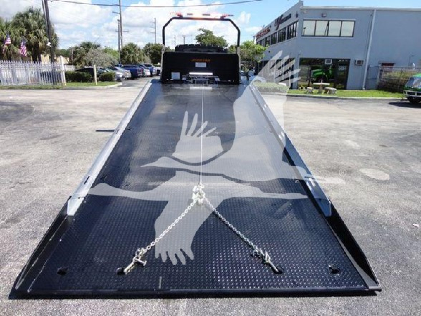 Truck rollback bed with black diamond-plate surface and central wheel chocks. View from rear toward cab. Safety chain anchors visible.