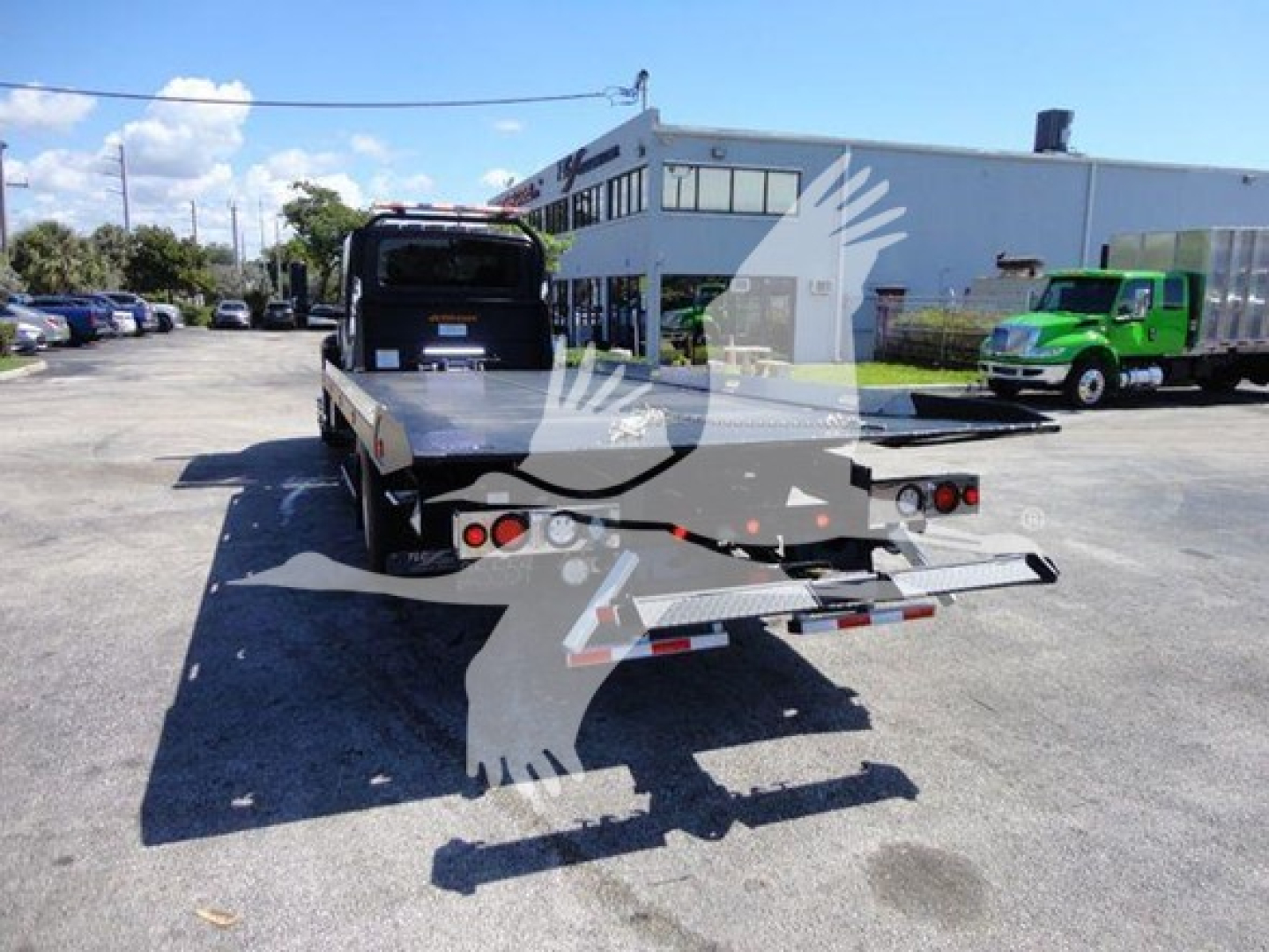 Truck rollback bed with rear view showing dual taillights, ramps, and reflective safety markings; yard with dealership building in background.