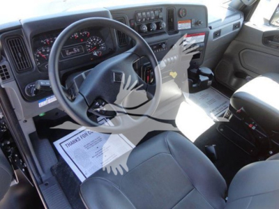 Interior of a 2022 International MV truck cab showing steering wheel, dashboard, and driver seat. Visible text includes VIN tag area and instrument cluster labels, along with a floor mat and paperwork on the seat.