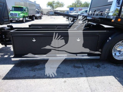 Truck rollback bed with storage toolbox and hydraulic tilt mechanism; side view showing black metal toolbox with two circular locks and chrome wheel; part of trailer hitch and chassis visible.