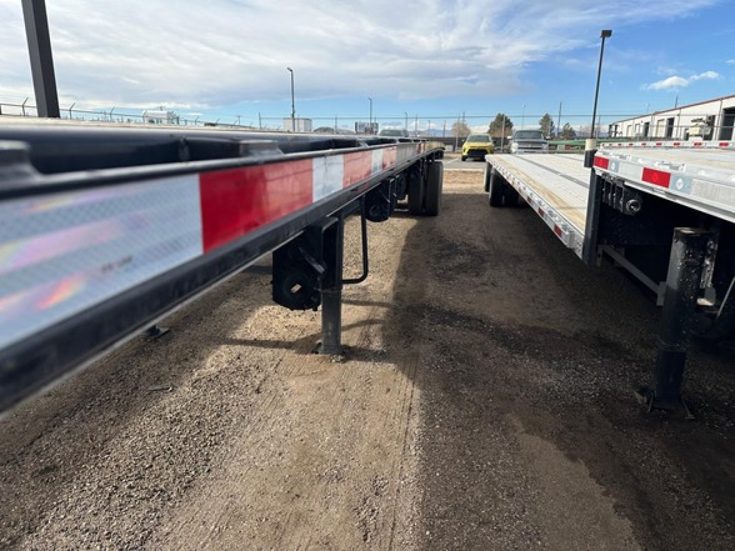 Trailer frame side with reflective red/white tape and support legs, long flatbed trailer in a lot, other trailers in background.